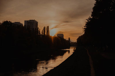 Silhouette buildings by river against sky at sunset