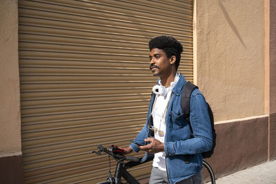 Afro man with bicycle holding smart phone by wall during sunny day