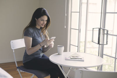 Woman text messaging on smart phone while having coffee at table by window in living room