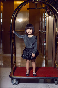 Beautiful korean girl child in a black dress is sitting on a luggage trolley in a hotel