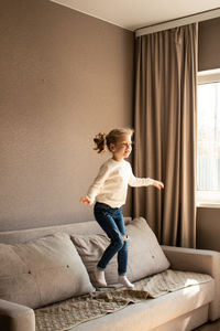 4 years old blonde girl joyfully jumps on couch at home, giggling with excitement. candid childhood 