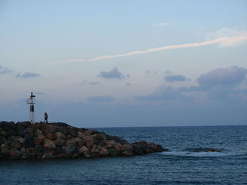 People standing on groyne in sea against sky during sunset