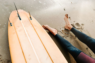 From above cropped anonymous surfer man dressed in wetsuit sitting with surfboard on the beach during sunrise