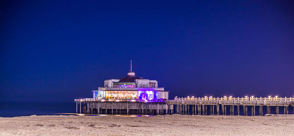 Illuminated building by sea against clear blue sky at night
