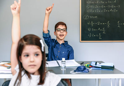 Students with mask on table raising their hands at school. selective focus on boy in background