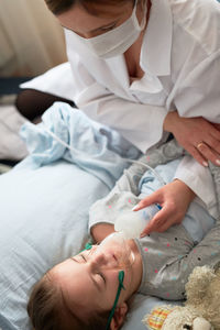 High angle view of female doctor wearing mask sitting by girl sleeping on bed