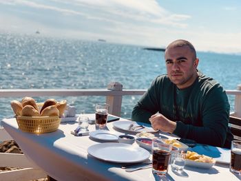 Portrait of man sitting on table by sea against sky