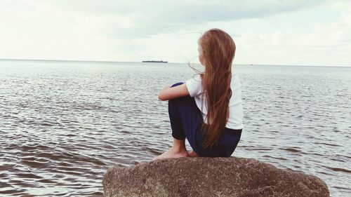 Rear view of girl sitting on rock at beach against sky