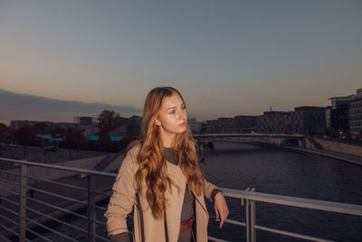 Beautiful woman looking away while leaning on railing against sky during sunset