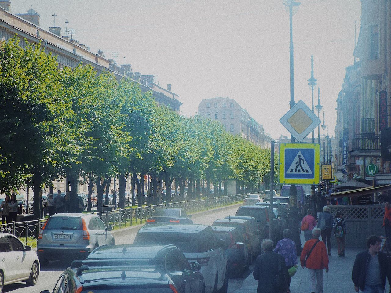 transportation, car, sign, motor vehicle, city, mode of transportation, road, tree, land vehicle, street, plant, communication, road sign, architecture, nature, traffic, group of people, real people, built structure, day, outdoors