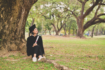 Smiling young woman in graduation gown holding certificate while sitting on grassy field at park