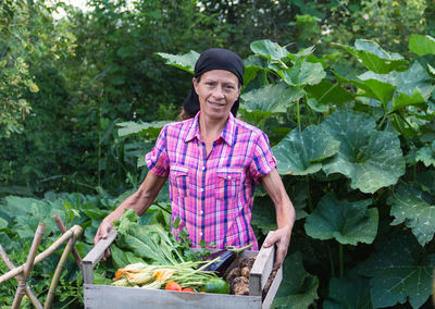 Portrait of farmer holding vegetables standing against trees