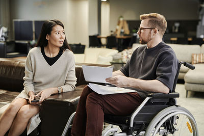Disabled colleague with documents talking to businesswoman sitting on sofa in office