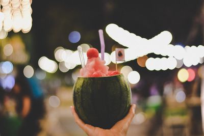 Cropped hand holding coconut against illuminated lights at night