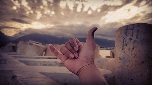 Cropped image of hand showing shaka sign against cloudy sky during sunset