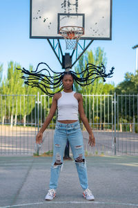 Full body young african american female in trendy ripped jeans and white top with waving braids standing against basketball hoop in urban park
