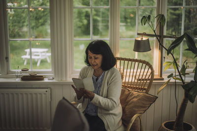 Mature woman using smart phone while sitting on chair near window at home