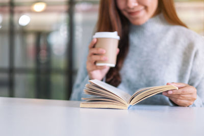 Midsection of woman drinking coffee while reading book on table