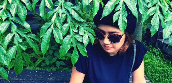 Portrait of young woman wearing sunglasses standing against plants and green leaves