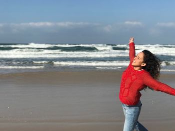 Happy woman with arms outstretched standing at beach against sky