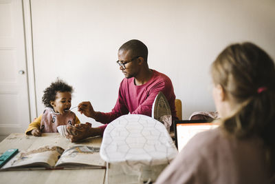 Father feeding daughter while mother working on laptop