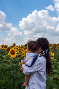 Mother holding boy with sunflower at field against sky