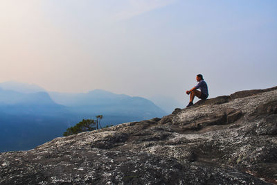 Side view of man sitting on cliff against sky