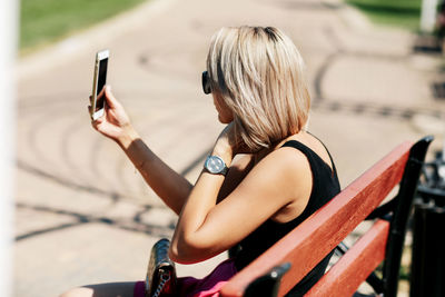 A young woman takes a selfie sitting on a park bench, adjusts her makeup and hairstyle