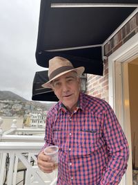 Portrait of man wearing hat holding drink standing against built structure on balcony 