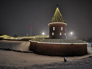 Illuminated lighthouse by building against sky at night