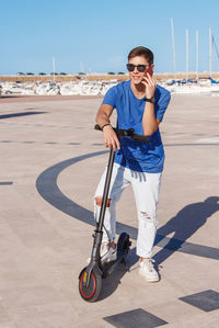 Young man stays with an electric scooter near the sea in the marina and talking on a smartphone