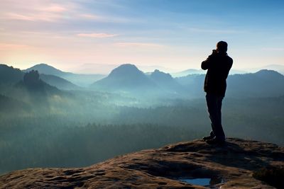 Photographer takes photos with big camera on peak of rock. dreamy misty landscape, hot sun above
