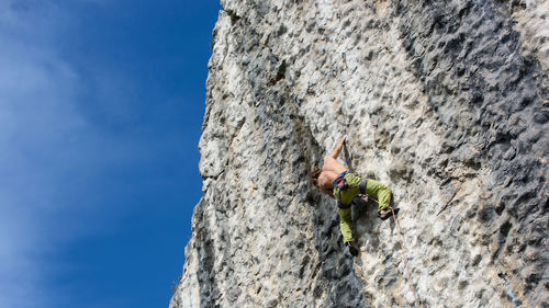 Low angle view of shirtless man climbing on rocky mountain against blue sky