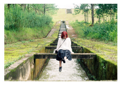 Woman sitting on wood over canal amidst field