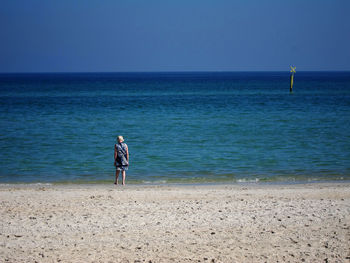 Rear view of woman standing at beach against clear blue sky during sunny day