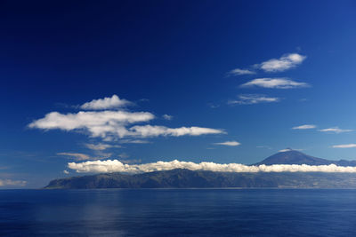 Scenic view of clouds over sea by mountain against blue sky