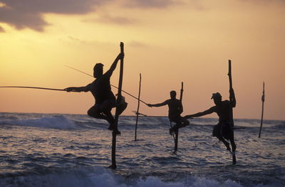 Men stilt fishing in sea against sky during sunset