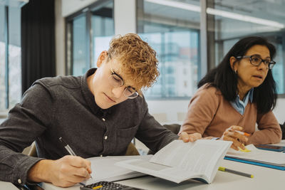 Blond man writing while sitting by mature female student at desk in classroom