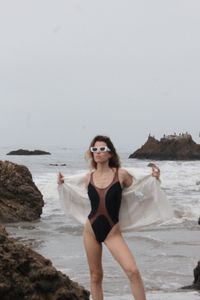 Portrait of young woman standing at beach against sky