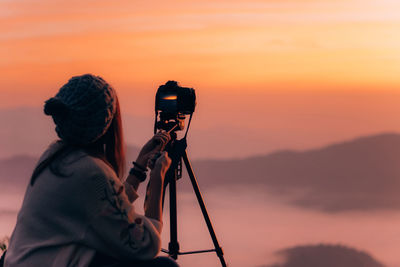 Side view of mid adult woman photographing while standing on mountain against sky during sunset