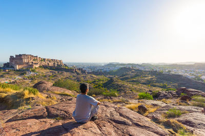 Rear view of man sitting on cliff against sky