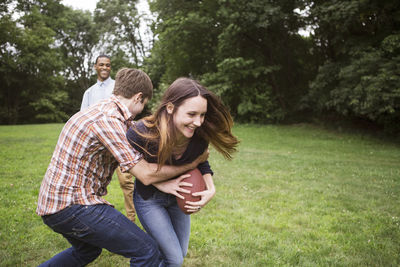 Man pulling back woman holding football ball while friend standing on grassy field