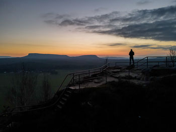 Silhouette person standing on railing against sky during sunset