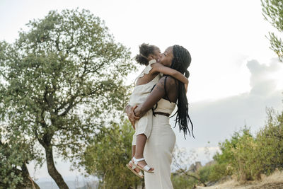 Side view of glad african american mom in white clothes with braided hair smiling and embracing daughter while standing against lush trees and cloudy sky in countryside