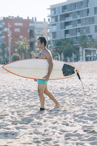 Full length of shirtless man carrying surfboard on beach