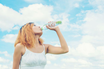 Young woman drinking water from bottle against cloudy sky