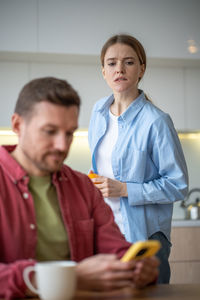 Woman peeping in man smartphone standing behind his back on kitchen at home.