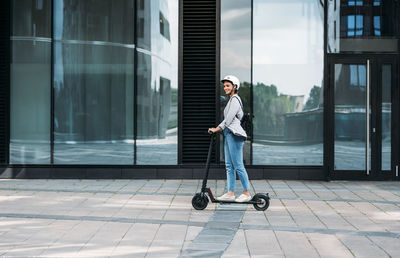 Young businesswoman wearing cycling helmet riding push scooter on footpath	
