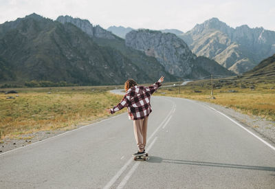 Young woman on skateboard on road against the beautiful mountain landscape, chemalskiy tract, altai