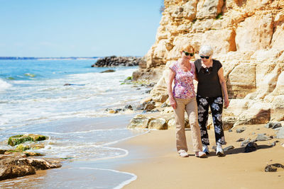 Two elderly women are walking along the rocky shore